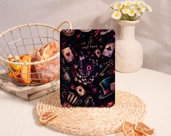 May include: A rectangular plaque featuring a dark, whimsical illustration with the text 'we all mad here'. The design includes playing cards, a top hat, a key, a potion bottle, and a teacup against a starry, purple and black background.