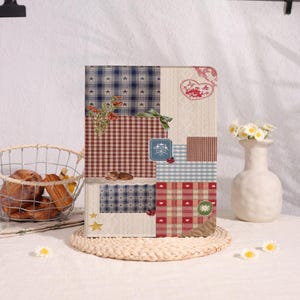 May include: A decorative notebook with a patchwork design featuring various patterns like stars, gingham, and hearts. The notebook has a neutral background and is adorned with a small floral arrangement and a white vase with daisies. A wire basket holds baked goods.