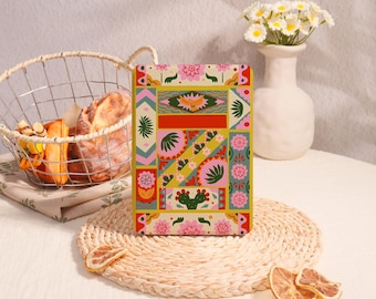 May include: A rectangular notebook with a floral and geometric design in pink, green, yellow, and red. A wire basket with bread and a vase with white flowers are in the background. The notebook is on a woven mat.