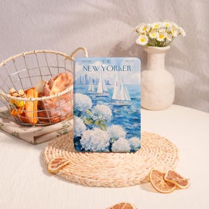 May include: A decorative rectangular item featuring a "The New Yorker" magazine cover with sailboats on the water and white flowers. The item is displayed on a woven placemat with dried orange slices, near a basket of bread and a vase of flowers.