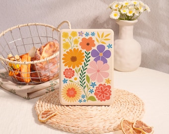 May include: An off-white rectangular case adorned with a colourful floral pattern, including orange, yellow, purple, and pink blooms. The case rests on a woven mat, with a bread basket and a vase of flowers in the background.