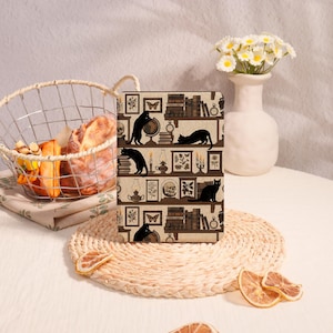 May include: A decorative journal featuring a repeating pattern of black cats, books, and vintage decor on a brown background. The design includes shelves with books, framed pictures, and a globe. The journal is displayed on a woven placemat.