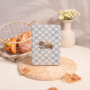 May include: A rectangular, blue and white checkered notebook featuring a dachshund illustration wearing a striped shirt and a floral crown. The notebook is placed on a woven placemat, with a basket of bread and a vase of flowers in the background.