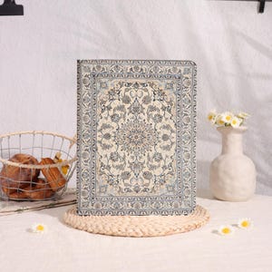 May include: A rectangular, cream-colored notebook or journal with a detailed, ornate rug-like pattern in shades of blue and beige. The notebook is propped up on a woven placemat, suggesting a home decor or stationery item.