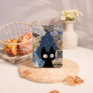 May include: A black cat illustration on a tablet case with a collage background featuring the word "Blue". The case is on a woven placemat, with dried orange slices and a basket of bread in the background.