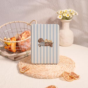 May include: A rectangular, light blue and white striped box featuring a watercolor illustration of a dachshund wearing a striped shirt and adorned with flowers. The image is set on a woven placemat with dried orange slices.