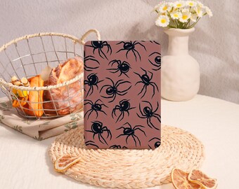 May include: A rectangular tablet case featuring a repeating pattern of black spiders on a dusty rose background. The case stands upright on a round woven mat, positioned near a wire basket filled with bread and a white vase with daisies.