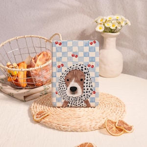 May include: A tablet case with a dog illustration wearing a polka-dot scarf, set against a blue and white checkered pattern with cherry details. The scene includes a bread basket, a vase of flowers, and a woven placemat.