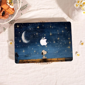 May include: Laptop skin featuring a night sky design with a crescent moon, stars, and a Snoopy character. The Apple logo is prominently displayed. The background is a deep blue with gold stars, and the Snoopy character is sitting on a brown rooftop.