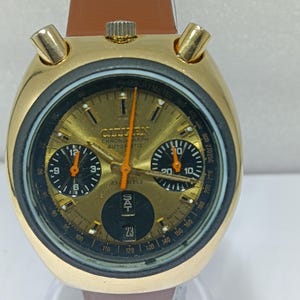 Peut inclure: Une montre automatique chronographe Citizen dorée avec un bracelet en cuir marron. Le cadran de la montre est doré avec des sous-cadrans noirs et des aiguilles orange. La date est affichée en bas. La montre a une échelle tachymétrique.