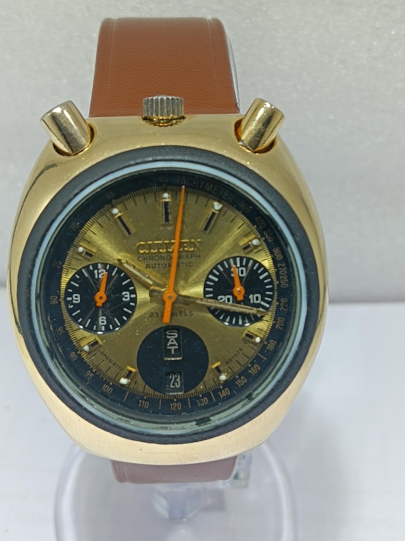 May include: A gold-toned Citizen chronograph automatic watch with a brown leather strap. The watch face is gold with black sub-dials and orange hands. The date is displayed at the bottom. The watch has a tachymeter scale.