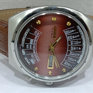 May include: A vintage Orient watch with a brown face and a stainless steel case. The watch features a day and date display, gold-colored hands and markers, and a brown leather band. The watch face displays "21 JEWELS" and "ORIENT".