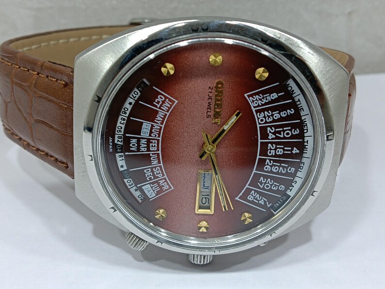 May include: A vintage Orient watch with a brown face and a stainless steel case. The watch features a day and date display, gold-colored hands and markers, and a brown leather band. The watch face displays "21 JEWELS" and "ORIENT".