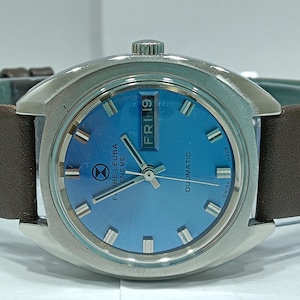 May include: A silver wristwatch with a blue face and a brown leather strap. The watch face has a date window that reads "FRI 19" and the text "DUDMATIC" and "F.RE.LEUBA GENEVE" printed on it.
