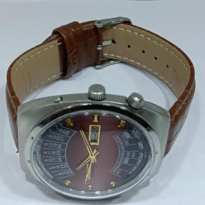 May include: A vintage wristwatch with a brown leather band and a silver-toned case. The watch face is burgundy with gold-colored hands and markers, and features a calendar function. The band is approximately 9 inches long.