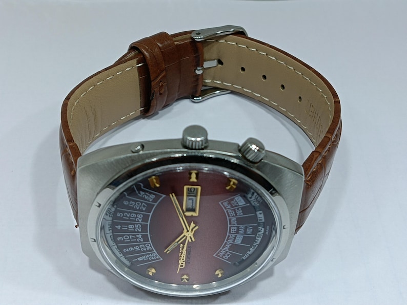 May include: A vintage wristwatch with a brown leather band and a silver-toned case. The watch face is burgundy with gold-colored hands and markers, and features a calendar function. The band is approximately 9 inches long.