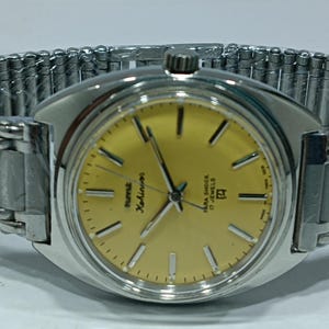 May include: A vintage wristwatch with a silver case and a yellow dial. The watch has black hour markers and hands, and a silver, expandable band. The dial includes the text "HMT Kohinoor" and "Para Shock 17 Jewels".