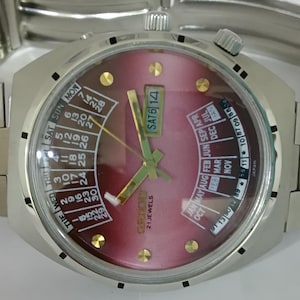 May include: A silver wristwatch with a red and white calendar face. The watch face has a gold-colored center and gold-colored hands. The watch face displays the day of the week, the date, and the month. The watch face also has the text "SAT 6 14" and "21 JEWELS" on it. The watch has a silver metal band.
