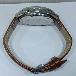 May include: A wristwatch with a silver-toned case and a brown leather strap. The watch has a round face with several buttons and a buckle closure. The strap is stitched with a light-colored thread.