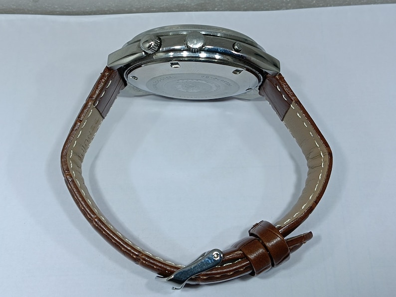 May include: A wristwatch with a silver-toned case and a brown leather strap. The watch has a round face with several buttons and a buckle closure. The strap is stitched with a light-colored thread.