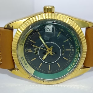 Puede incluir: Un reloj de pulsera dorado con una esfera negra y un anillo exterior verde. El reloj tiene números arábigos y la palabra "Satellite" impresa en la esfera. El reloj tiene una correa de cuero marrón.