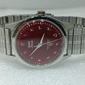 May include: A silver wristwatch with a burgundy face and Arabic numerals. The watch features the text "hmt Janata" and "Para Shock 17 Jewels." The watch has a silver, expandable band.