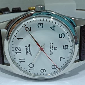 May include: A silver-toned wristwatch with a white face, black numerals, and red second hand. The watch has a brown leather band and the text "HMT Janata" and "Para Shock 17 Jewels" on the face. The watch is made in India.