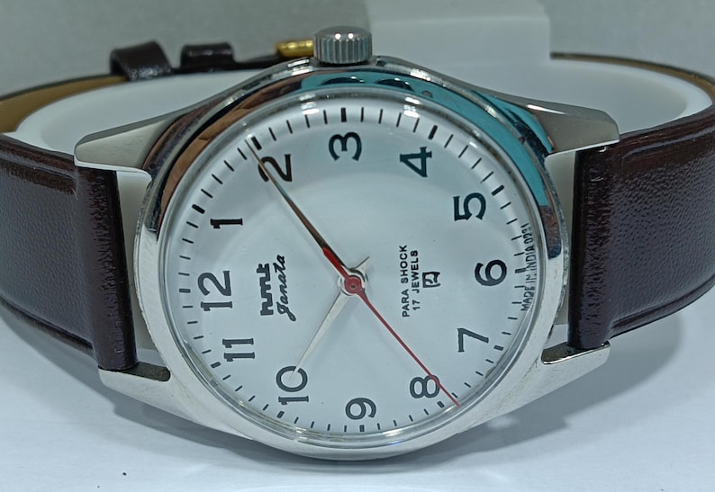 May include: A silver-toned wristwatch with a white face, black numerals, and red second hand. The watch has a brown leather band and the text "HMT Janata" and "Para Shock 17 Jewels" on the face. The watch is made in India.
