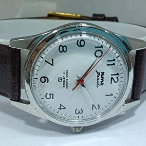 May include: Silver-tone analog watch with a white face, black numerals, and red second hand. The watch has a brown leather band and the text "hmt Janata" is visible on the face. The watch is a classic style.