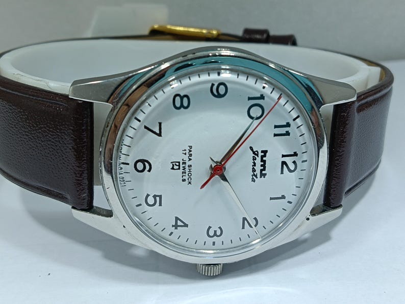 May include: Silver-tone analog watch with a white face, black numerals, and red second hand. The watch has a brown leather band and the text "hmt Janata" is visible on the face. The watch is a classic style.