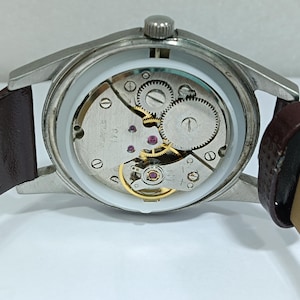 May include: Close-up of a vintage wristwatch with a silver-colored case and a brown leather strap. The watch's inner workings are visible, showcasing gears, jewels, and other mechanical components. The watch is displayed against a white background.