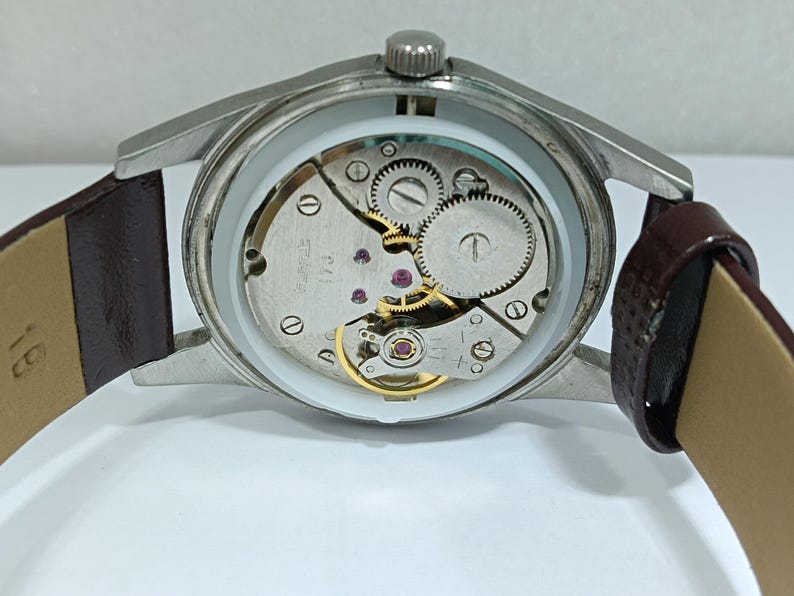 May include: Close-up of a vintage wristwatch with a silver-colored case and a brown leather strap. The watch's inner workings are visible, showcasing gears, jewels, and other mechanical components. The watch is displayed against a white background.