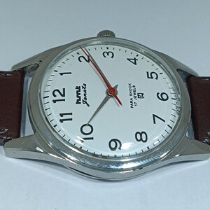 May include: Close-up of a silver-toned wristwatch with a white face, black numerals, and red second hand. The watch has a brown leather strap. The watch face reads "hmt Janata" and "Para Shock 17 Jewels".
