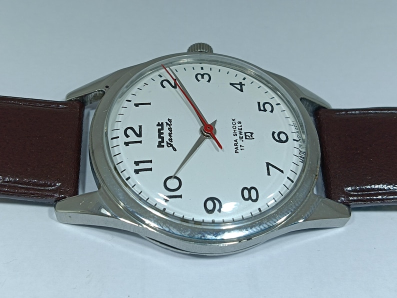 May include: Close-up of a silver-toned wristwatch with a white face, black numerals, and red second hand. The watch has a brown leather strap. The watch face reads "hmt Janata" and "Para Shock 17 Jewels".