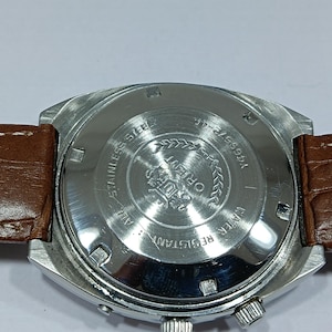 May include: A stainless steel Orient watch with a brown leather strap. The watch face is silver with black text and a silver bezel. The back of the watch has the words "Stainless Steel" and "Water Resistant".