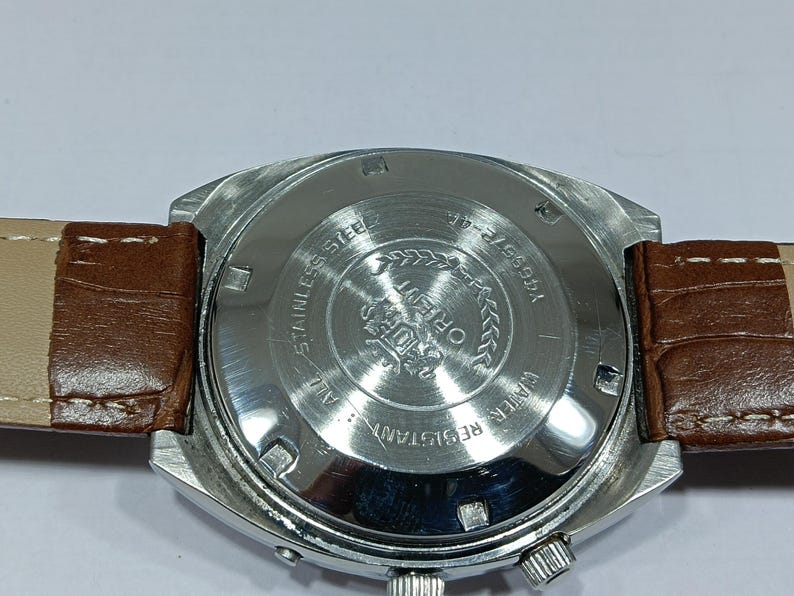 May include: A stainless steel Orient watch with a brown leather strap. The watch face is silver with black text and a silver bezel. The back of the watch has the words "Stainless Steel" and "Water Resistant".