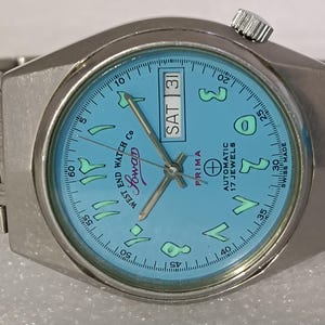 May include: A silver wristwatch with a light blue face. The watch has Arabic numerals, a day-date display, and the text "WEST END WATCH Co Swan PRIMA AUTOMATIC 17 JEWELS SWISS MADE". The watch band is silver.