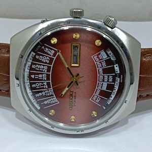 May include: A vintage Orient watch with a brown face, silver case, and brown leather band. The watch features a day and date display, along with a calendar function. The watch face displays the days of the week and months.