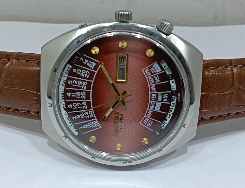 May include: A vintage Orient watch with a brown face, silver case, and brown leather band. The watch features a day and date display, along with a calendar function. The watch face displays the days of the week and months.