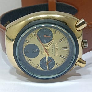 May include: A gold-toned Citizen chronograph automatic watch with a brown leather strap. The watch face is gold with black sub-dials and hands. The day and date are displayed. The watch has a retro design.