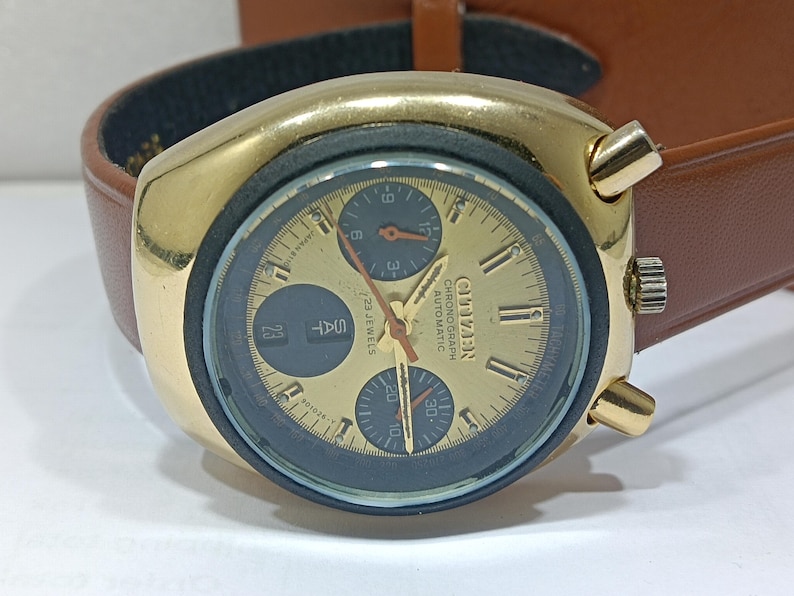 May include: A gold-toned Citizen chronograph automatic watch with a brown leather strap. The watch face is gold with black sub-dials and hands. The day and date are displayed. The watch has a retro design.
