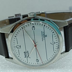 Könnte beinhalten: Eine silberne Armbanduhr mit weißem Zifferblatt und schwarzen Zeigern. Die Uhr hat ein braunes Lederarmband und auf dem Zifferblatt steht "HMT Janata Para Shock 17 Jewels Made in India".