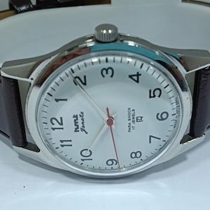 May include: Silver-toned wristwatch with a white face, black numerals, and a red second hand. The watch features a brown leather band and the text "hmt Janata" on the face. The watch has a classic, minimalist design.