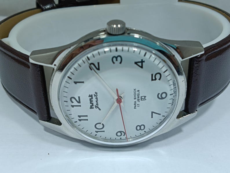 May include: Silver-toned wristwatch with a white face, black numerals, and a red second hand. The watch features a brown leather band and the text "hmt Janata" on the face. The watch has a classic, minimalist design.