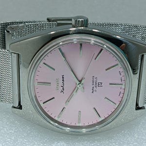 Pode incluir: Um relógio de pulso prateado com um mostrador rosa e uma pulseira de malha prateada. O mostrador do relógio tem o texto "Seiko" e "Harlequin" impresso nele. O relógio também tem o texto "Para Shock 17 Jewels" impresso no mostrador.