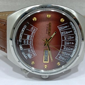 May include: A vintage Orient watch with a brown face and a calendar function. The watch has a silver-colored case, gold-colored hands and markers, and a brown leather band. The watch face displays the day, date, and month.