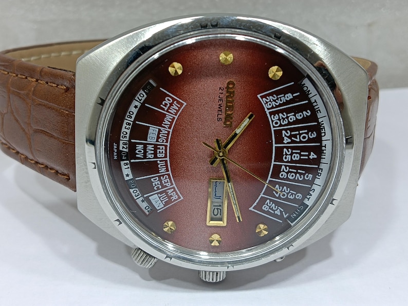 May include: A vintage Orient watch with a brown face and a calendar function. The watch has a silver-colored case, gold-colored hands and markers, and a brown leather band. The watch face displays the day, date, and month.