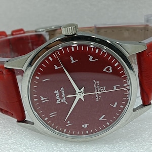 May include: A wristwatch with a burgundy dial and a matching leather strap. The watch has silver hands and hour markers. The dial features Arabic numerals and the brand name "hmt Janata".