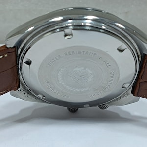 May include: Close-up of a vintage wristwatch with a brown leather band. The watch has a silver-colored stainless steel case and a white dial with the text "WATER RESISTANT" and "ALL STAINLESS STEEL". The model number is Y469672-4A.