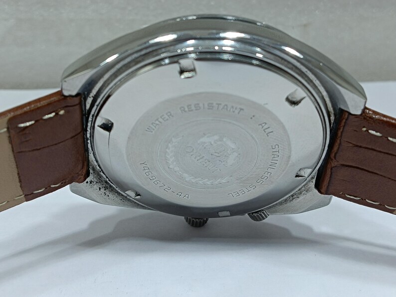 May include: Close-up of a vintage wristwatch with a brown leather band. The watch has a silver-colored stainless steel case and a white dial with the text "WATER RESISTANT" and "ALL STAINLESS STEEL". The model number is Y469672-4A.